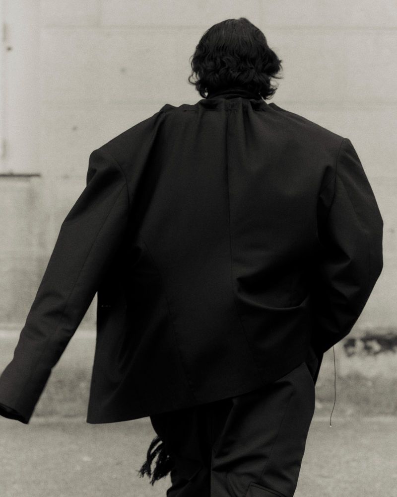 Person from behind wearing black jacket and pants on city street. Back view of individual in dark fashion attire in urban environment.