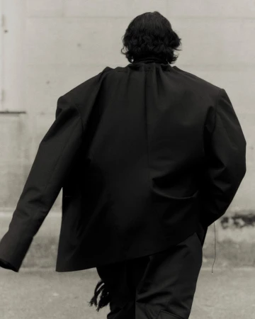 Person from behind wearing black jacket and pants on city street. Back view of individual in dark fashion attire in urban environment.