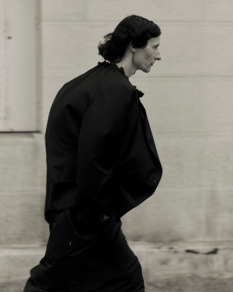 Monochrome profile of an androgynous person wearing a black ruffled collar, standing in front of a wall.
