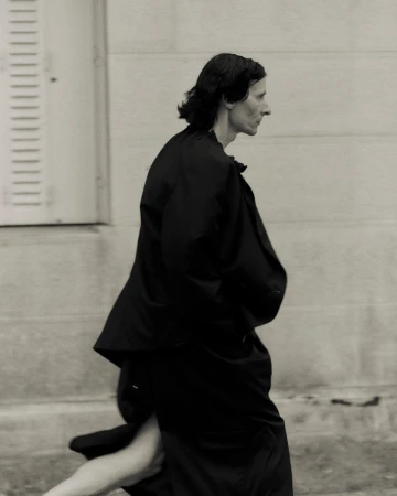 Monochrome fashion shot of a slender adult woman in profile, wearing a dark overcoat and moving near a light-colored building with louvered doors.