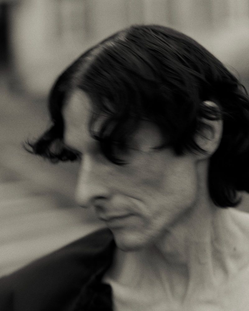 Monochrome portrait of an adult male with dark, wavy hair, seen in profile against a blurred light background, wearing a dark open jacket.