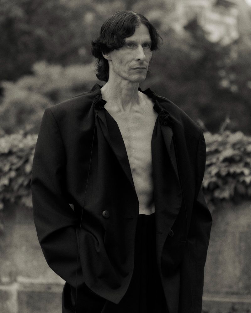 Monochrome portrait of a slender adult male in an oversized black blazer, posed outdoors against a backdrop of dense green foliage.