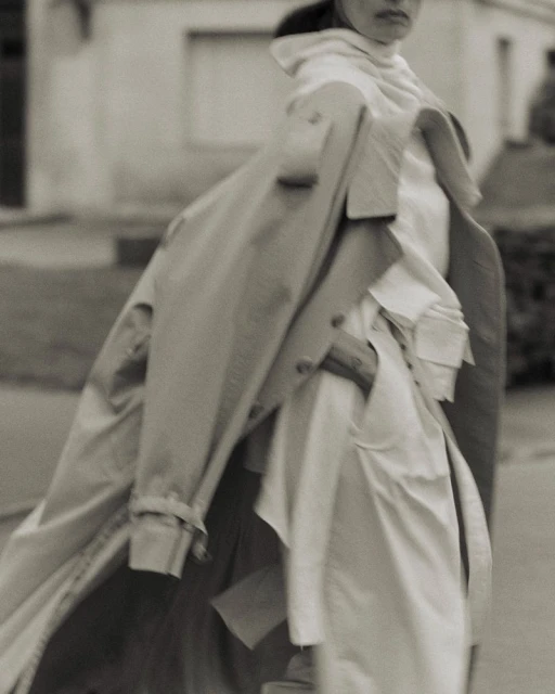 Sepia portrait of a woman walking in an urban environment, wearing a trench coat over a white outfit on a sidewalk with residential buildings.