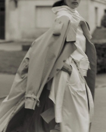 Sepia portrait of a woman walking in an urban environment, wearing a trench coat over a white outfit on a sidewalk with residential buildings.