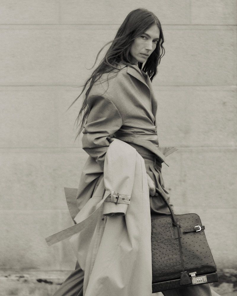 Monochrome portrait of an adult female model in a trench coat, holding a structured handbag, with hair blowing in the wind.