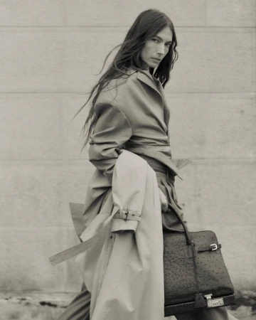 Monochrome portrait of an adult female model in a trench coat, holding a structured handbag, with hair blowing in the wind.