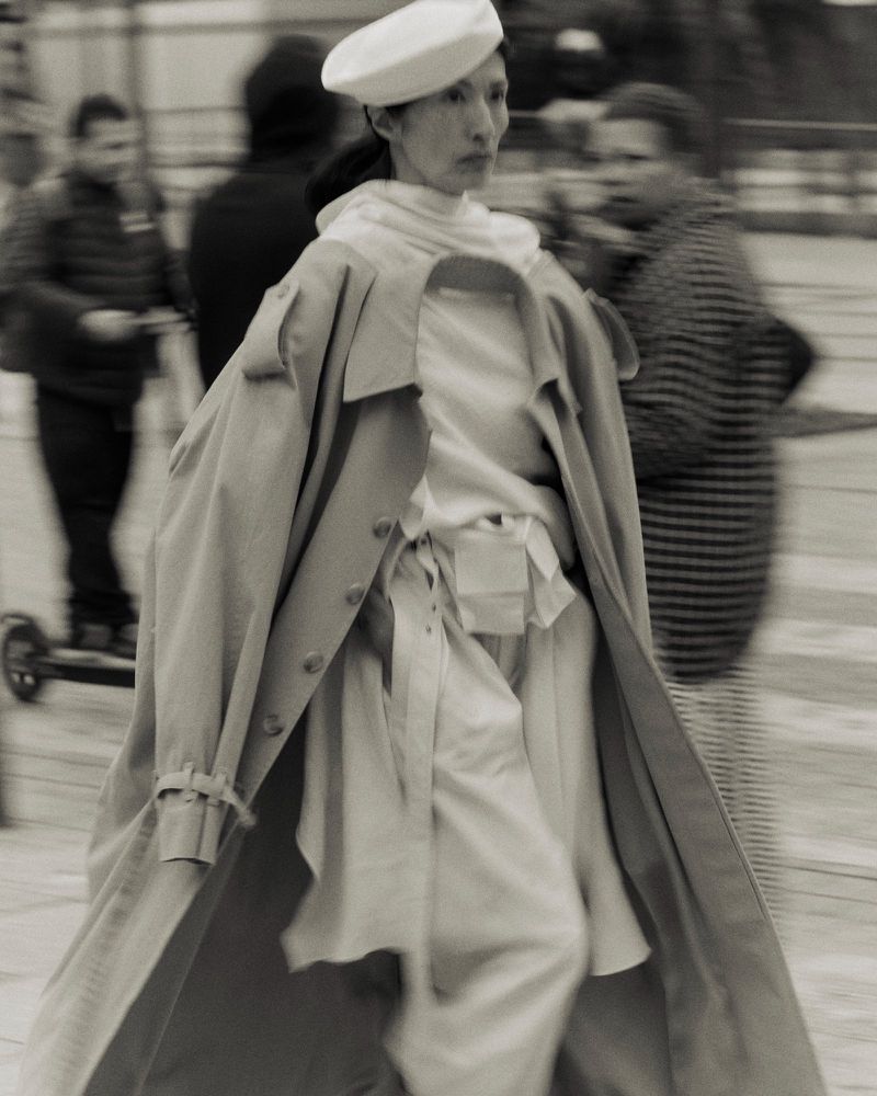 Monochrome image of an adult female walking in a neutral trench coat and a white beret, with blurred people in the background.
