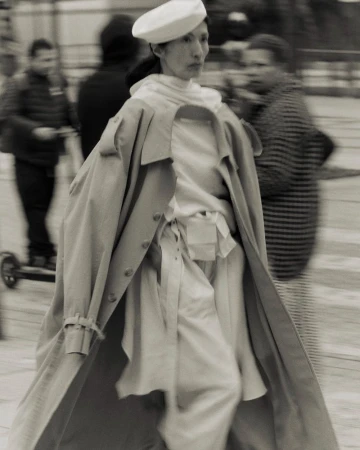 Monochrome image of an adult female walking in a neutral trench coat and a white beret, with blurred people in the background.