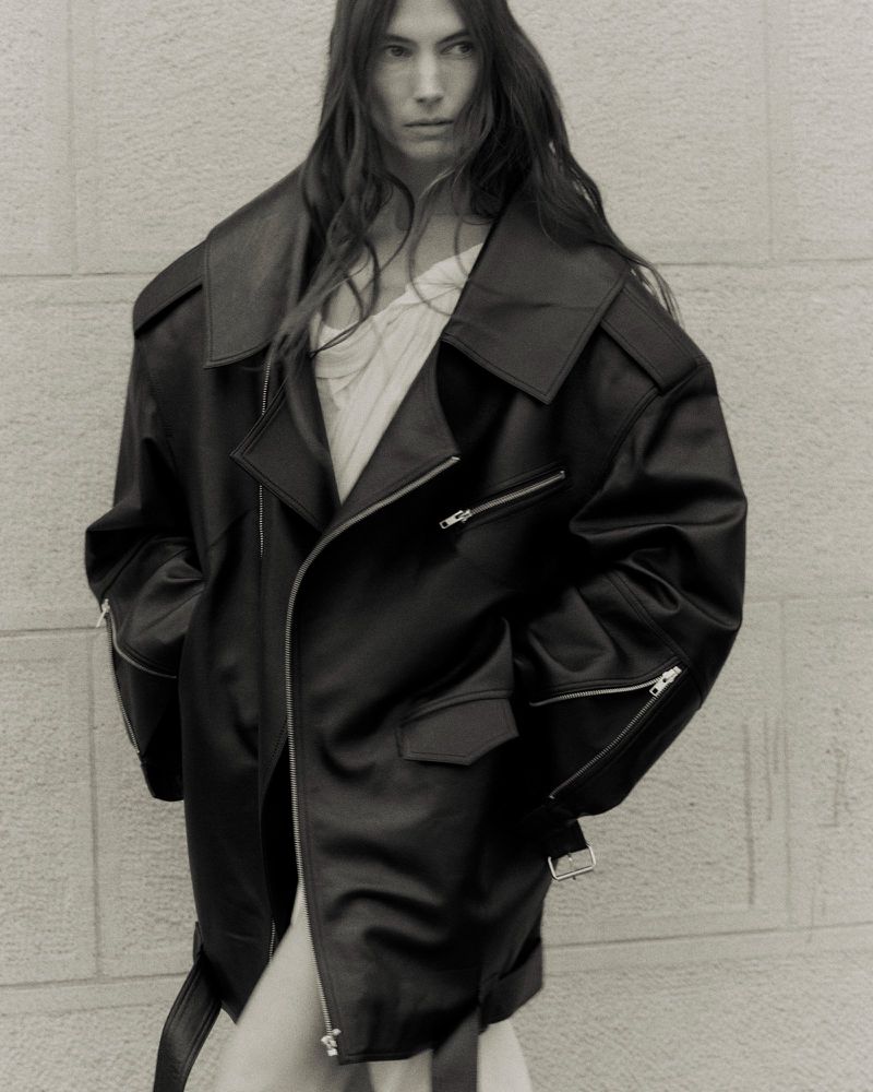 Woman with long hair poses in an oversized black leather moto jacket in front of a textured wall in a fashion editorial shot.