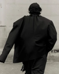 Model seen from behind wearing an oversized black blazer standing outdoors against a pale wall for Vautrait