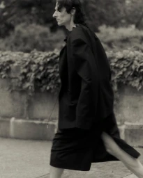 Model in a black jacket and skirt walking outdoors past a low stone wall for Vautrait