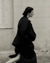 Model walking in a black jacket and skirt against a building exterior with a shuttered window