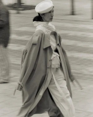Model wearing a white pillbox hat and long coat walking on an outdoor paved street in a campaign shot by Dudi Hasson for Vautrait