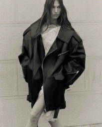 Model wearing an oversized black leather jacket and white dress against a stone wall for Vautrait campaign.