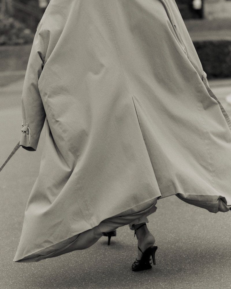 Model wearing a long beige trench coat and heeled sandals walking outdoors on asphalt