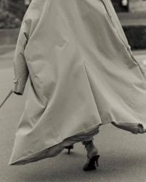 Model wearing a long beige trench coat and heeled sandals walking outdoors on asphalt