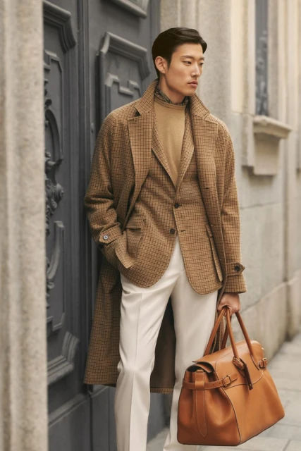 Asian man in a brown plaid overcoat and cream pants holds a tan leather duffel bag, standing near an ornate grey doorway outdoors.