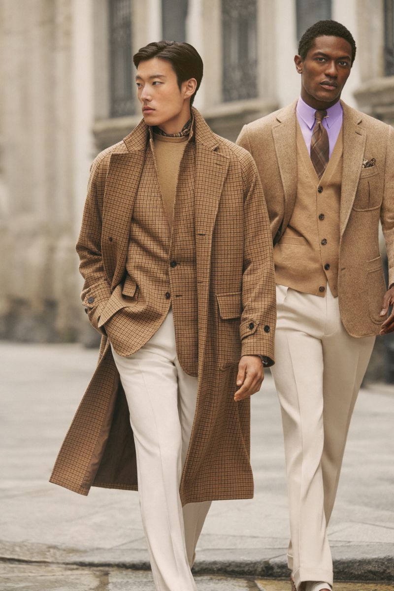 Two male fashion models showcase tan and white menswear on a city street. One wears a tan coat, the other a suit and vest.