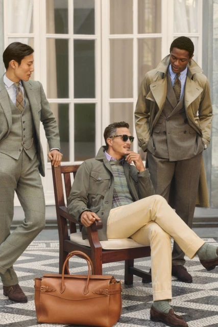 Three men in classic style attire pose outdoors; one seated in olive jacket and tan pants, flanked by two in patterned suits and coats.