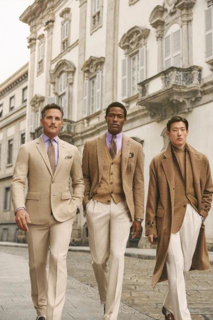 Three well-dressed men in suits walking on a city street with ornate buildings, showcasing fashion and style.