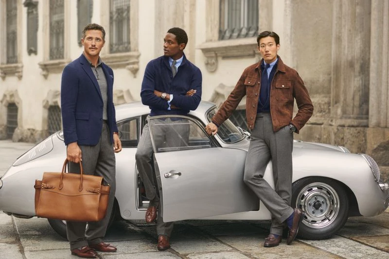 Three well-dressed men pose next to a silver classic car outdoors. One carries a brown leather briefcase in an urban setting.