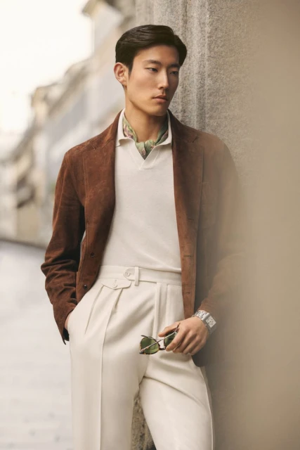 Adult male in a brown jacket and cream-colored pants stands outdoors in a medium shot, holding sunglasses, wearing a watch and neck scarf.