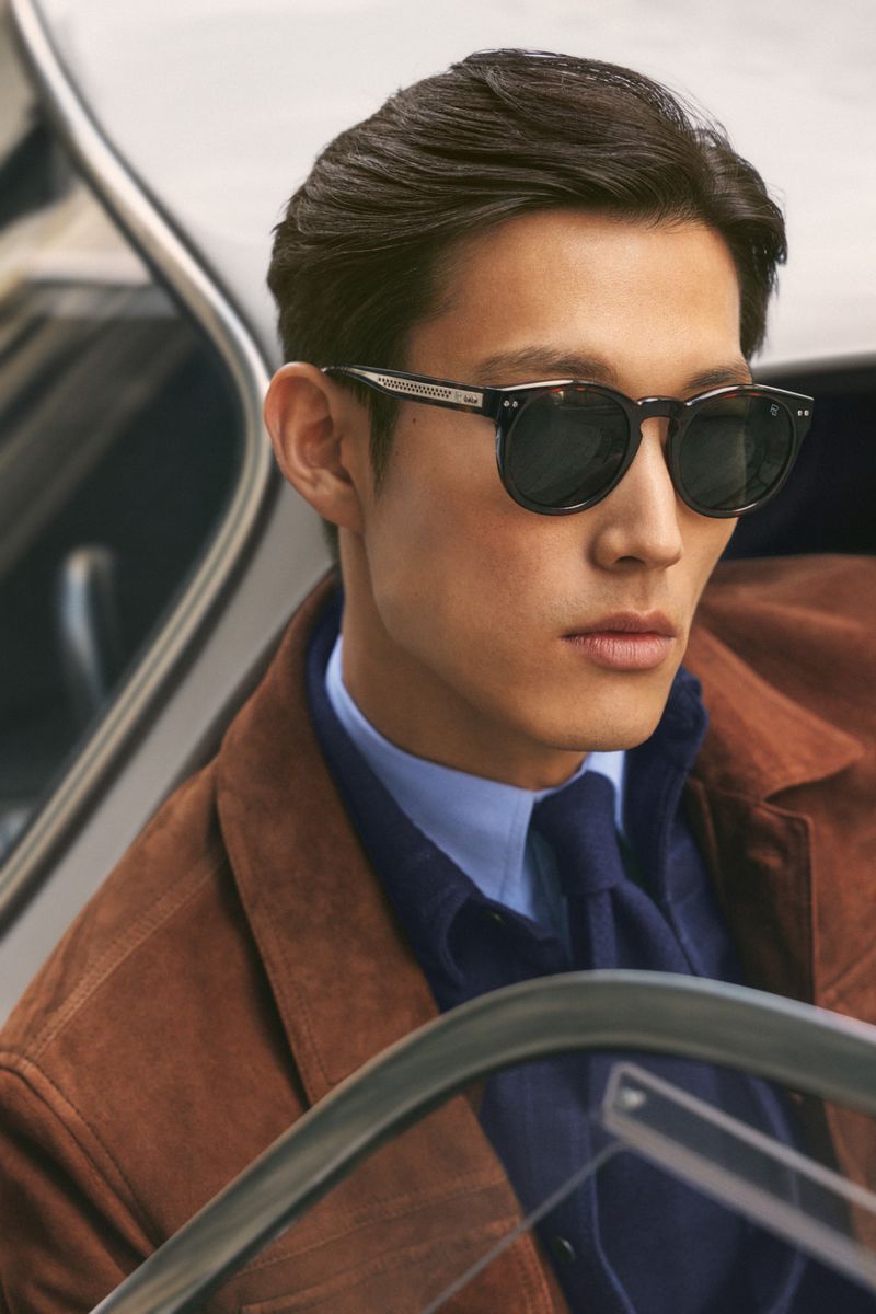A close-up studio shot features a stylish man with dark hair wearing sunglasses, a brown jacket, and a blue shirt and tie, exuding confidence.