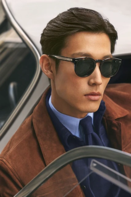 A close-up studio shot features a stylish man with dark hair wearing sunglasses, a brown jacket, and a blue shirt and tie, exuding confidence.