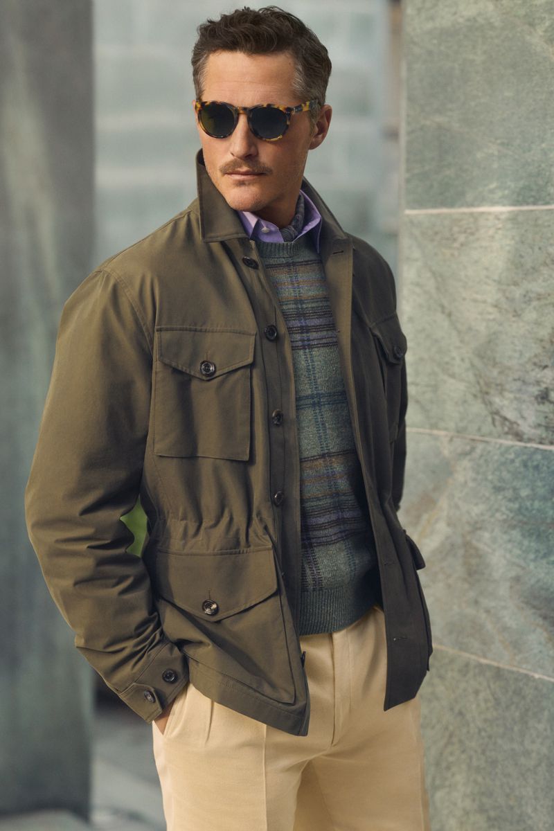 Adult male in sunglasses wearing a green jacket and khaki pants. He is indoors against a gray and green column.