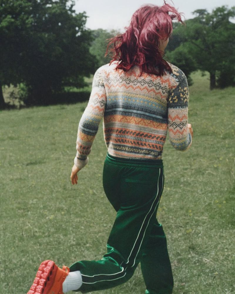 Woman runs through sunlit meadow. Back view of person with colorful sweater and sweatpants jogging on grassy field with trees.