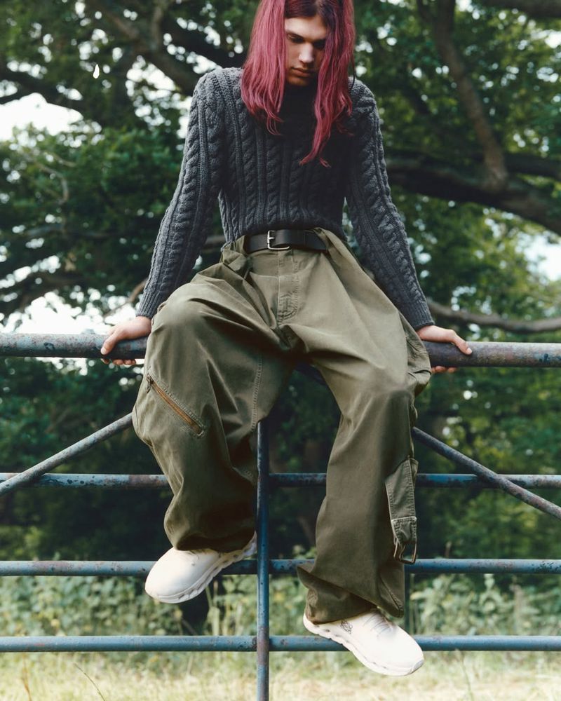 A young adult with pink hair sits on a metal fence wearing an grey sweater and oversized green cargo pants in a grassy, outdoor setting.