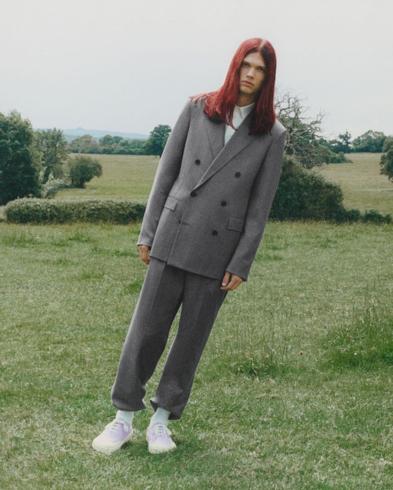 Model with long red hair wearing a grey double-breasted suit standing in an open grassy field