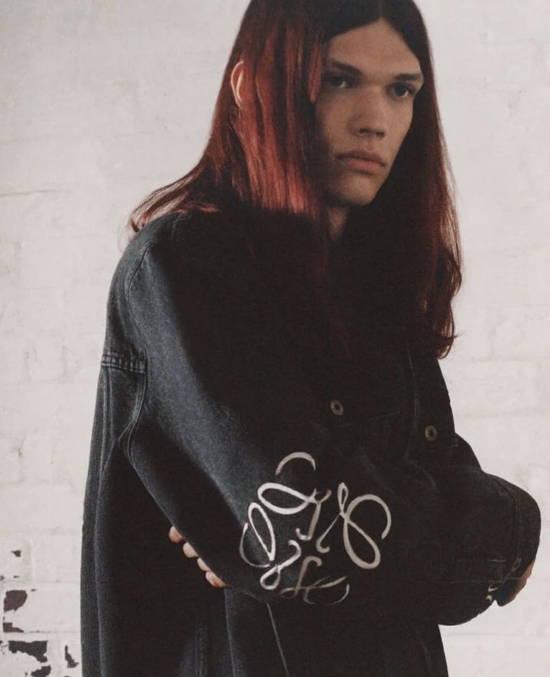 Portrait of a long-haired male model in a dark denim jacket with embroidered logo on the sleeve, set against a white brick wall.