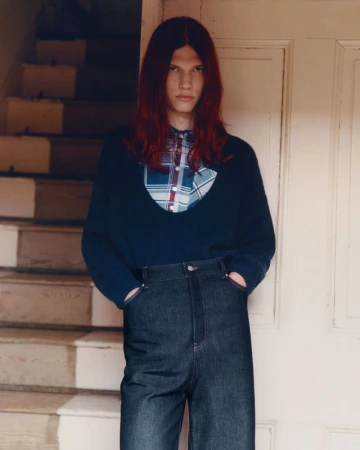 A young adult poses in a blue sweater and jeans against a door, with stairs in the background, indoors with natural lighting.