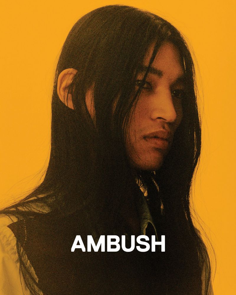 Portrait of a person with long dark hair and a light complexion against a yellow backdrop, featuring the text 'AMBUSH' in white.