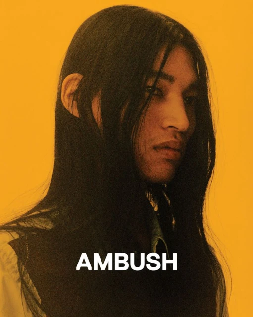 Portrait of a person with long dark hair and a light complexion against a yellow backdrop, featuring the text 'AMBUSH' in white.