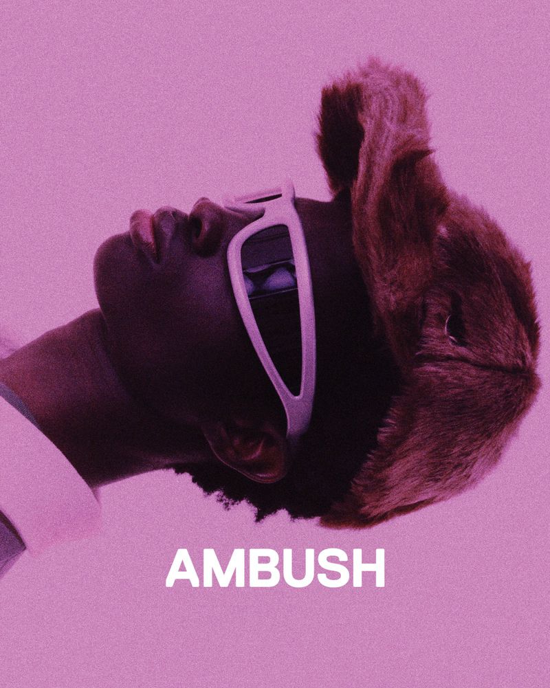 Portrait of a person wearing sunglasses and a fur hat against a pink background, featuring the brand name AMBUSH in white letters.