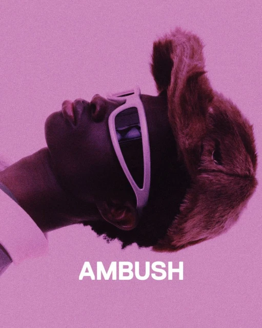 Portrait of a person wearing sunglasses and a fur hat against a pink background, featuring the brand name AMBUSH in white letters.
