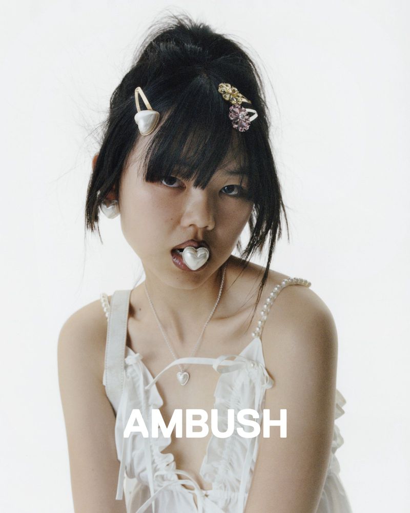 Close-up portrait of a young woman with dark hair and fair skin, wearing a white top and heart jewelry, set against a bright white background.