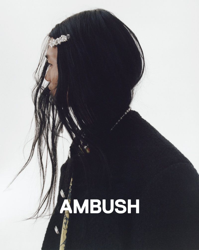 Profile of a person with long dark hair wearing a black jacket and a beaded necklace against a white background, and a hair accessory.