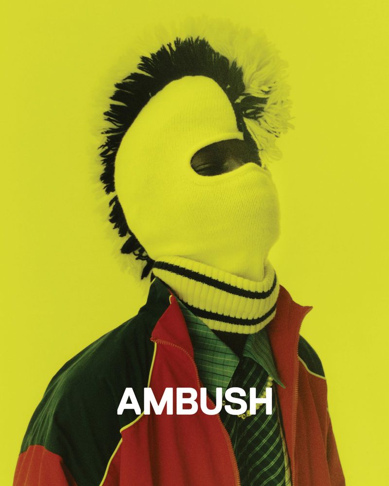 Person wearing a yellow balaclava with a black mohawk, striped scarf, and red jacket against a vibrant yellow background.
