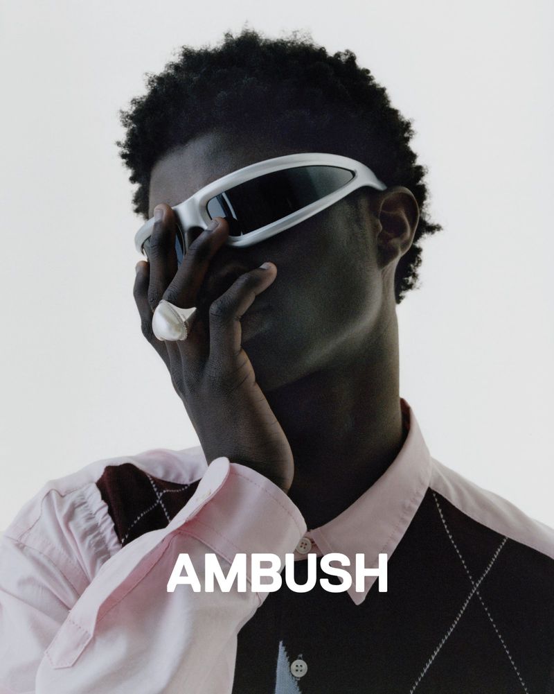 An adult male model wearing a pink shirt, sunglasses, and a ring poses in front of a white backdrop; the 'AMBUSH' logo is displayed at the bottom.