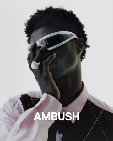 An adult male model wearing a pink shirt, sunglasses, and a ring poses in front of a white backdrop; the 'AMBUSH' logo is displayed at the bottom.