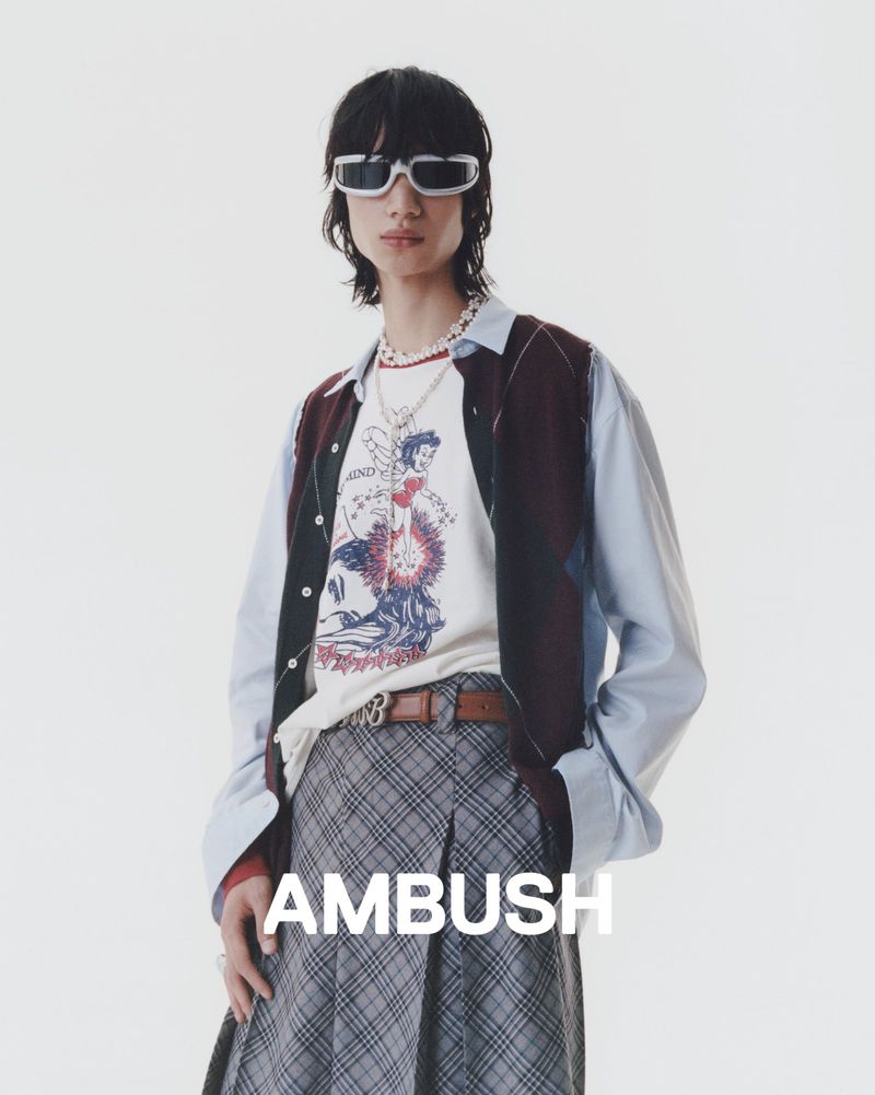 Studio shot of a young adult male wearing sunglasses, graphic t-shirt, patterned skirt, and light blue button-down shirt.