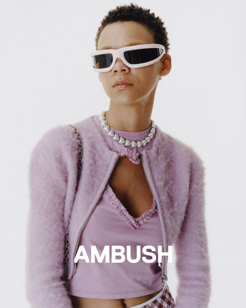 Stylish woman in pink fuzzy jacket, tank top, and white sunglasses, with a pearl necklace, poses against a bright white backdrop.