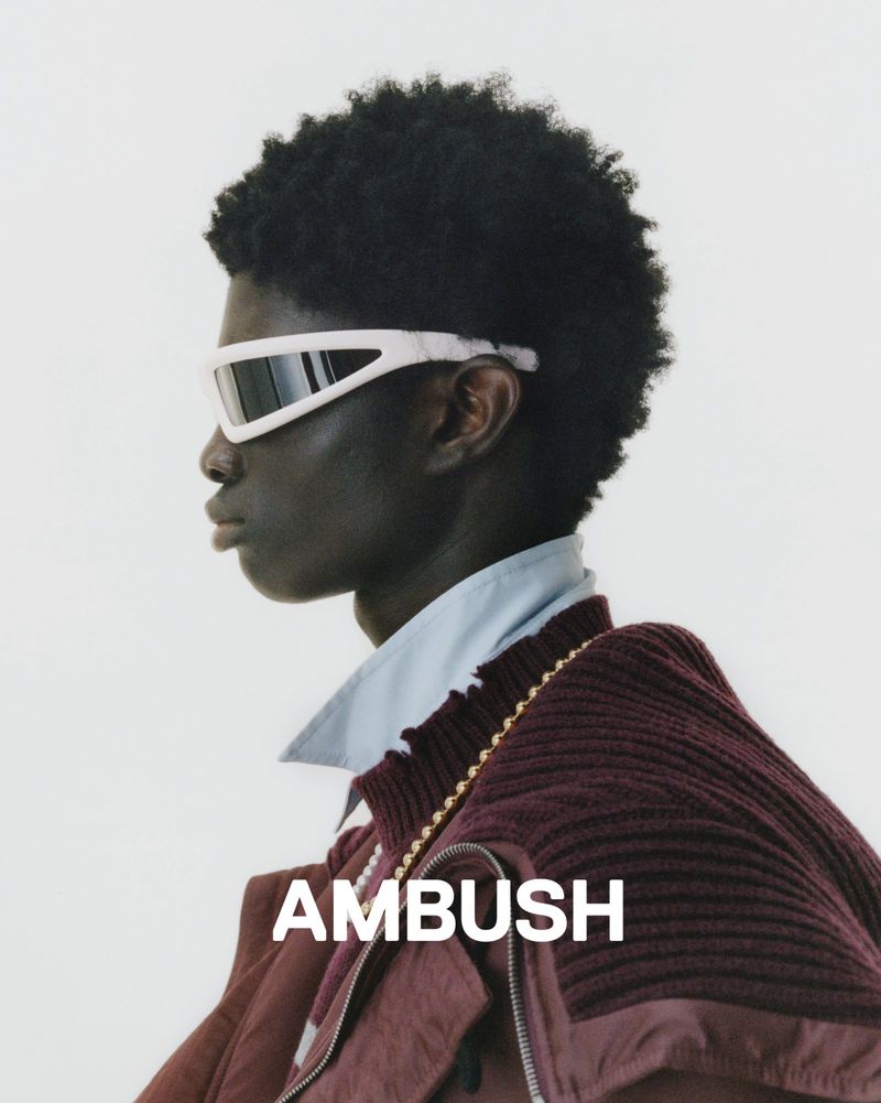 Profile shot of a person with dark skin, wearing sunglasses, a collared shirt, and a jacket against a white background. Brand logo visible.