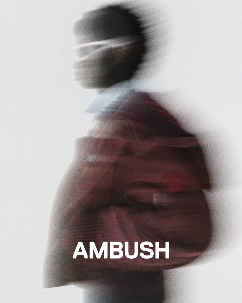 Blurred motion fashion shot featuring an adult with a maroon jacket against a bright white background with the word 'AMBUSH' in the foreground.
