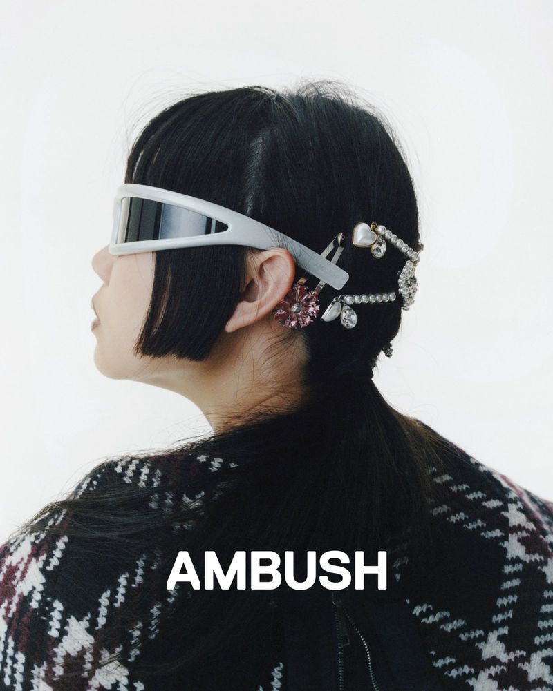 A woman with dark hair, wearing futuristic visor glasses and decorative hairpins with a black and white patterned jacket, poses in a studio.