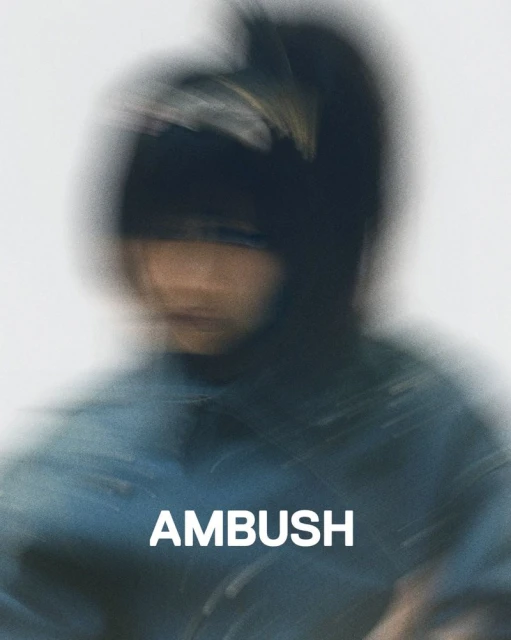 Close-up shot of a blurry person wearing a dark beanie and blue top against a white background with the brand name AMBUSH in white letters.
