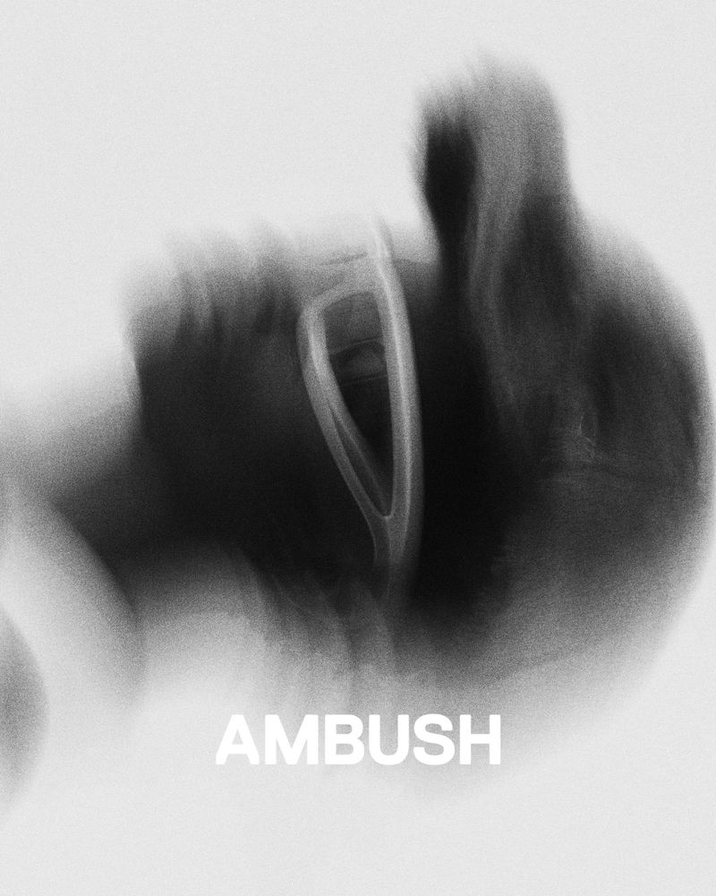 Monochromatic abstract blurry image of a bent metal wire against a gradient white and gray background with the text AMBUSH at the bottom.
